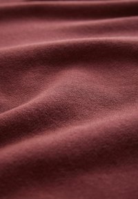 Textilie v hluboké burgundské barvě s jemnou, hladkou texturou, která má jemné vrásky a záhyby, jež zdůrazňují hloubku látky.