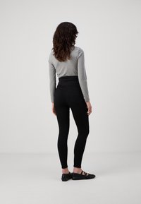 Mujer con cabello rizado de espaldas, vistiendo una camiseta de manga larga gris, pantalones negros de talle alto y zapatos planos negros, contra un fondo liso.