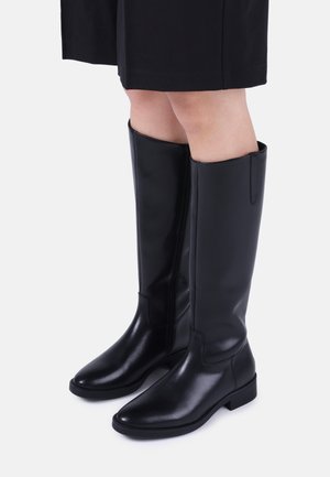 Bottes en cuir noires arrivant aux genoux avec des talons bas, portées avec une jupe noire sur un fond blanc uni.