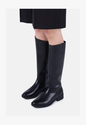 Bottes en cuir noires arrivant aux genoux avec des talons bas, portées avec une jupe noire sur un fond blanc uni.