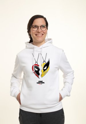 Usměvavá žena s brýlemi, která nosí bílou mikinu s přívěsky červené a žluté masky Deadpool a nápisem "Come Together Marvel Studios."