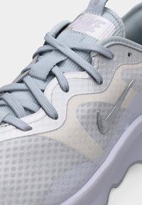 Κοντινό πλάνο ανοιχτόγριου αθλητικού παπουτσιού Nike με υφή υφάσματος, χονδρές κορδόνια, μεταλλικό λογότυπο swoosh και μαξιλαρωμένη γλώσσα.