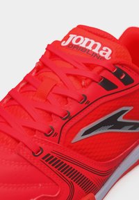 Φωτεινό κόκκινο αθλητικό παπούτσι με κορδόνια, μαύρο και ασημί λογότυπο στο πλάι και ένδειξη "joma DRIBLING" στη γλώσσα.