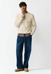 Beiger Pullover mit Grafikdesign, darüber ein braunes kariertes Hemd getragen. Weite blaue Jeans aus Denim, kombiniert mit hellbraunen Wildleder-Loafers.