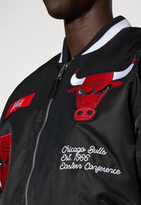 Fekete bomber dzseki vörös Chicago Bulls logóval, fehér csíkozott gallérral és hímzett szöveggel: "Chicago Bulls Alapítás éve 1966, Keleti Konferencia."