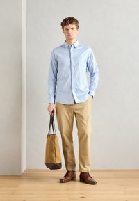 Chemise boutonnée bleu clair à manches longues, pantalons beiges, chaussures en cuir marron et sac fourre-tout marron et beige. Design simple, tissu texturé.