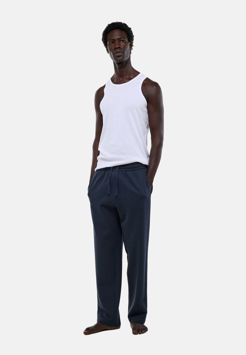 Débardeur sans manches blanc et pantalon de jogging bleu marine foncé avec une taille élastique et un cordon de serrage. Le mannequin est pieds nus, les mains dans les poches.