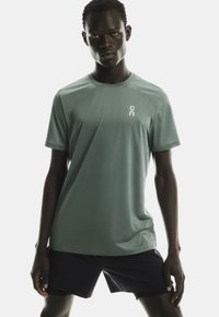 Model masculin îmbrăcat cu un tricou sport cu mânecă scurtă de culoare verde deschis și pantaloni scurți negri, stând cu picioarele depărtate pe un fundal alb simplu.