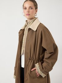 Ženska v rjavi jakni z bež karirastim notranjim podlogom in ovratnikom, ena roka v žepu, gleda rahlo vstran z nevtralnim izrazom.