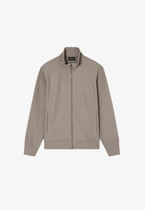 Vaaleanruskea vetoketjullinen takki, jossa pystykaulus, pitkät hihat ja kaksi etutaskua, esitetty valkoisella taustalla.