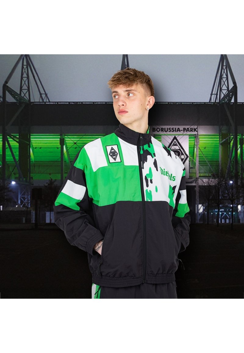 Zwarte en groene windbreaker jas met een ritssluiting aan de voorkant, een kleurblok ontwerp, witte accenten en een logo patch op de bovenste linkerborst.