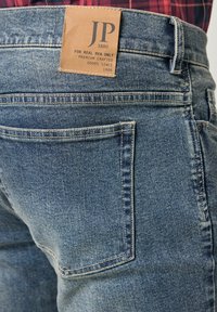 Pantalones vaqueros de mezclilla en un color azul desgastado, con una etiqueta de cuero marrón en la cintura que incluye la marca "JP" y detalles de costura.