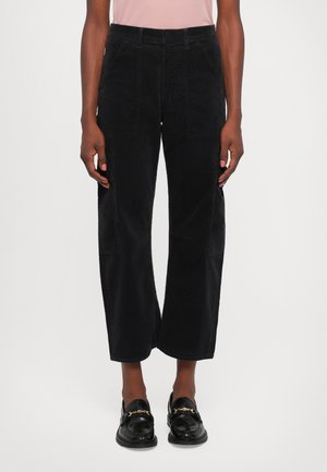 Person wearing black wide-leg pants with front and side pockets and black slip-on loafers with gold chain detail.