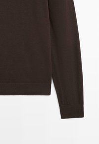 Mørkebrunt langærmet sweaterærme og kant med ribstrikkede manchetter mod en hvid baggrund.