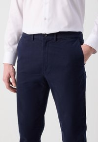 Man som bär mörkblå byxor med händerna i fickorna och en instoppad vit knäppt skjorta mot en enkel bakgrund.