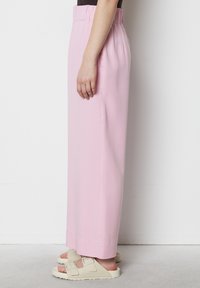 Pantaloni palazzo rosa chiaro con vita elastica arricciata, texture liscia e tasche laterali, abbinati a sandali beige.