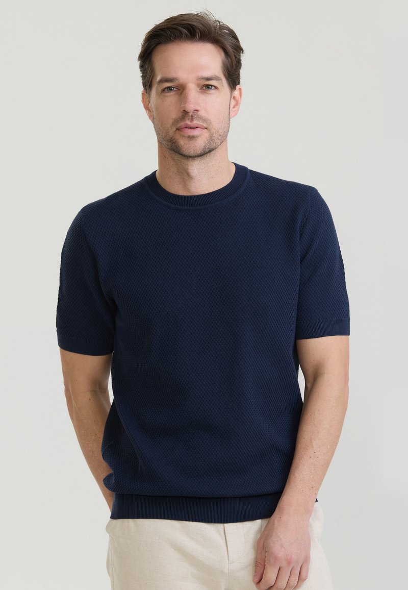 Erwachsener Mann mit kurzen braunen Haaren, der einen marineblauen strukturierten Kurzarmpullover und hellbeige Hosen trägt, stehend vor einem schlichten hellen Hintergrund.