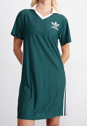 Kobieta w ciemnozielonej sukience Adidas z cienkimi pionowymi paskami, białym kołnierzykiem i logo Adidas na lewej piersi.