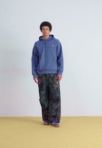Blue hoodie with a kangaroo pocket and drawstring; green camo pants; purple sneakers; standing against a plain background.