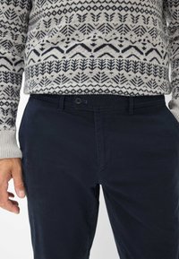 Pantaloni di cotone blu navy con chiusura a bottone, abbinati a un maglione grigio in maglia con un motivo geometrico nero.