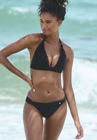 Černé bikini se trojúhelníkovým topem a nízko střiženými kalhotkami, vyrobené z hladké látky s jemným žebrováním. Model pózuje u vody.
