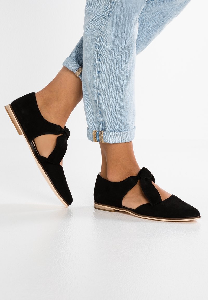Ballerines en daim noir avec un design découpé, un accent noué à l'avant et une semelle de couleur claire. Portées avec un jean en denim clair roulé.