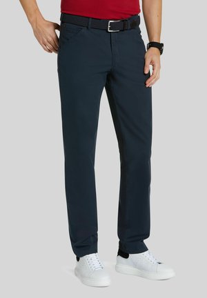 Homme portant un pantalon bleu marine, des baskets blanches, une ceinture noire tressée et une chemise rouge, debout avec les mains près des poches sur un fond uni.