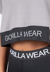 Grijze cropped T-shirt met zwarte band met "GORILLA WEAR" in het grijs. Zwarte shorts met witte "GORILLA WEAR" tailleband. Stof lijkt glad.
