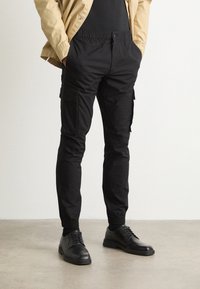 Czarne spodnie typu cargo z elastycznym pasem, wyposażone w liczne boczne kieszenie, zwężane nogawki oraz mankiety przy kostkach, zestawione z czarnymi butami.