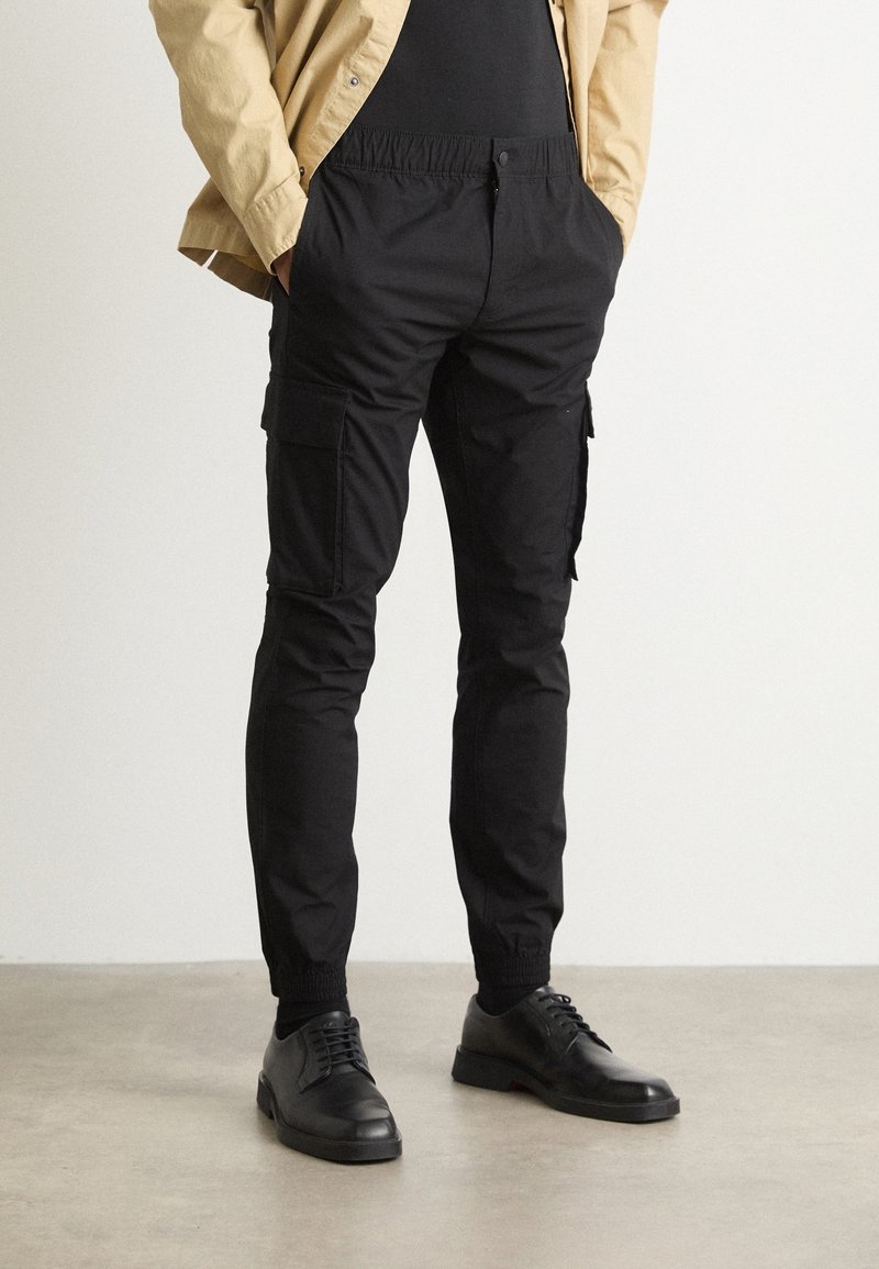 Czarne spodnie typu cargo z elastycznym pasem, wyposażone w liczne boczne kieszenie, zwężane nogawki oraz mankiety przy kostkach, zestawione z czarnymi butami.