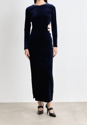 Vrouw gekleed in een navy fluwelen jurk met lange mouwen en een lengte tot de vloer, met uitsparingen en strikdetails aan de rechterkant, gecombineerd met zwarte hoge hakken.