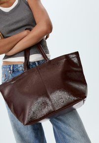 Person in einem grauen Tanktop und Jeans hält eine große, glänzende, dunkelbraune Ledertasche mit Doppelhöhen.