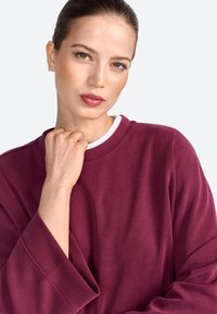 Felpa bordeaux con collo a giro e maniche lunghe. Il tessuto sembra morbido con una consistenza liscia. Presenta dettagli bianchi all'interno del colletto.