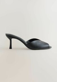 Mule en cuir noir à bout ouvert avec un talon moyen fin et une tige lisse et incurvée sur un fond clair uni.