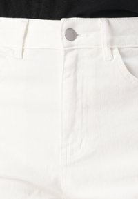 Pantalones vaqueros de mezclilla blanca con una cinturilla alta, que presentan un botón plateado, bolsillos cosidos y una tela suave y texturizada.