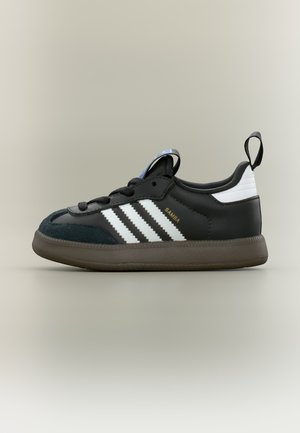 Μαύρο αθλητικό παπούτσι Adidas Samba με λευκές ρίγες, καφέ σόλα, μαύρα κορδόνια, λευκή ετικέτα στη φτέρνα και θηλιά στο πίσω μέρος.