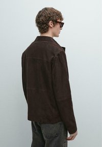 Giacca in camoscio marrone con colletto classico, dettaglio sulla spalla posteriore e taglio dritto, abbinata a jeans scuri, vista da dietro.