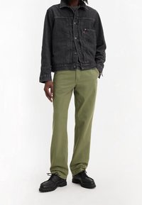 Chaqueta de mezclilla negra con botones plateados, pantalones verdes y zapatos negros de cordones con suelas texturizadas. Conjunto casual y en capas.