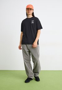 T-shirt en coton noir avec une petite impression graphique sur la poche, associé à un pantalon en velours côtelé gris et des baskets noires. Une casquette orange complète le look.