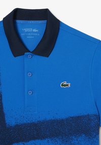 Polo bleu Lacoste Sport avec col noir, trois boutons, motif noir moucheté et logo crocodile vert sur la poitrine.