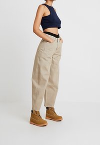 Námornícky crop top, béžové kalhoty s vysokým pasem a světle hnědé kotníkové boty s černými tkaničkami a šedými detaily, vystavené ve vzpřímené poloze.