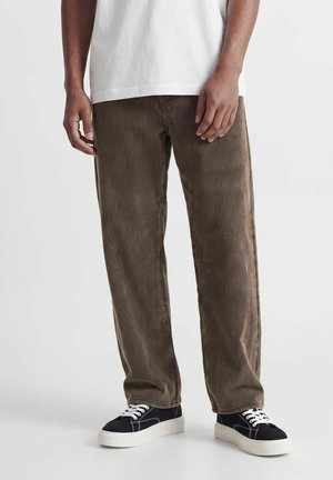 Personne portant un pantalon en velours côtelé marron, des baskets noires avec semelles et lacets blancs, et une chemise blanche, debout devant un fond uni.