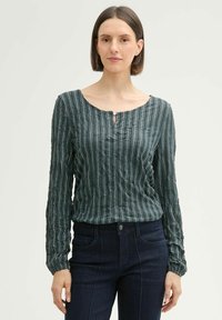 Dunkelgrüne Bluse mit strukturierten Streifen, Rundhalsausschnitt und Knopfdetail mit Schlitz. Kombiniert mit dunkelblauen Jeans und langen Ärmeln, die an den Manschetten gerafft sind.