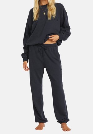 Kobieta ubrana w czarną, oversize'ową bluzę i dopasowane czarne spodnie dresowe ze ściągaczem, na bosaka, na jednolitym tle.