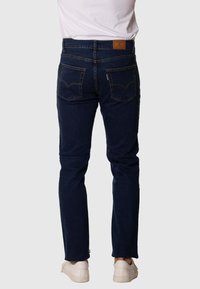 Jeans de mezclilla de un azul oscuro con corte ajustado, dos bolsillos traseros, costuras en contraste y una etiqueta de marca de cuero marrón en la cinturilla.