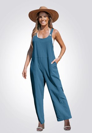 Blauwe linnen jumpsuit met wijde pijpen, verstelbare banden, een voorkantzak en houten knopenaccenten. Gecombineerd met een strohoed en beige sandalen.