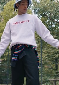 Weißes Sweatshirt mit rotem "carhartt."-Logo, schwarze Hose und ein mehrfarbig gestreifter Pullover um die Taille gebunden. Lässiger urbaner Stil.
