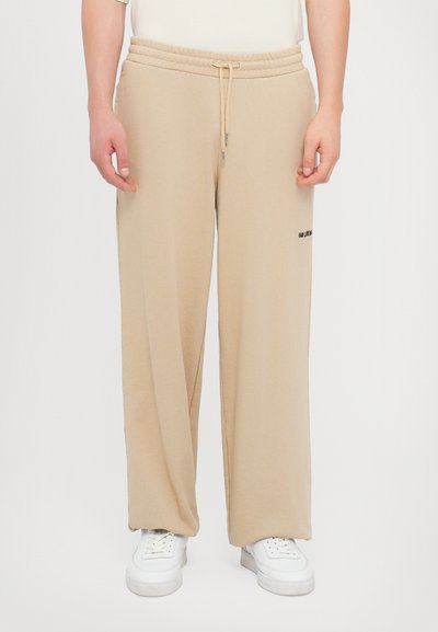 Beige sweatpants with a drawstring waist, straight fit, side pockets, and black logo detail on the right leg. Soft, textured fabric.