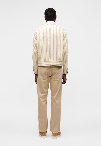 Homme portant une veste beige rayée, un pantalon beige et des chaussures beige, debout, dos tourné, sur un fond blanc uni.