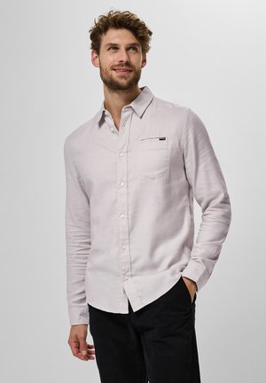 Camisa de manga larga rosa claro en tela texturizada con cuello abotonado, bolsillo en el pecho y etiqueta de marca sutil. Combinada con pantalones negros.
